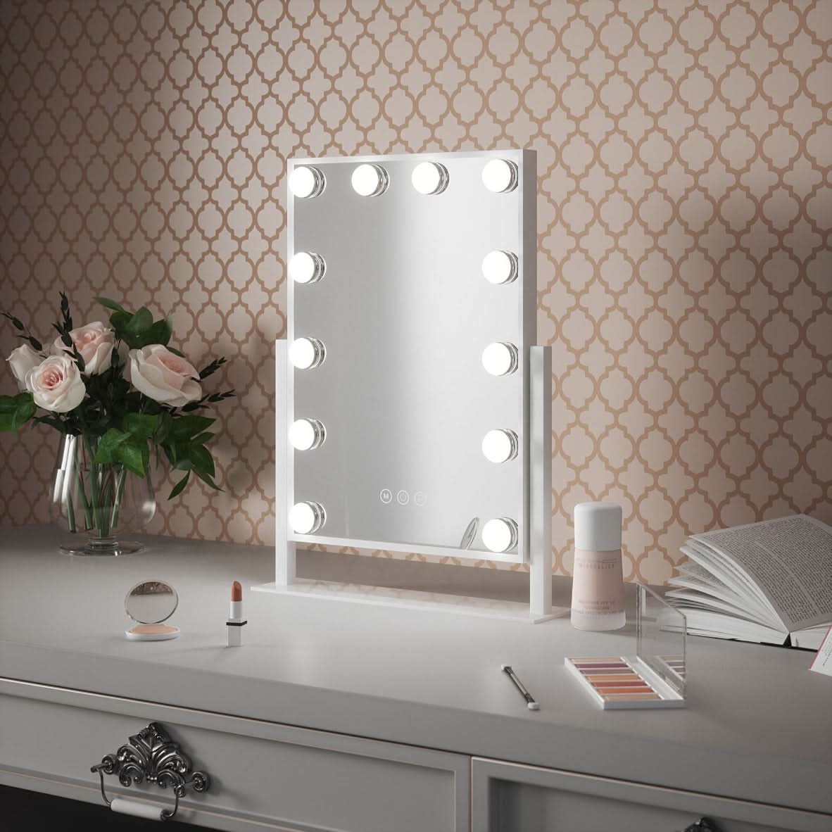 The Olsen Hollywood Mirror White 47x37cm by hollywood mirrors sits on a white vanity with adjustable LED bulbs, makeup items, a lipstick, flowers in a vase, and an open book against patterned wallpaper.